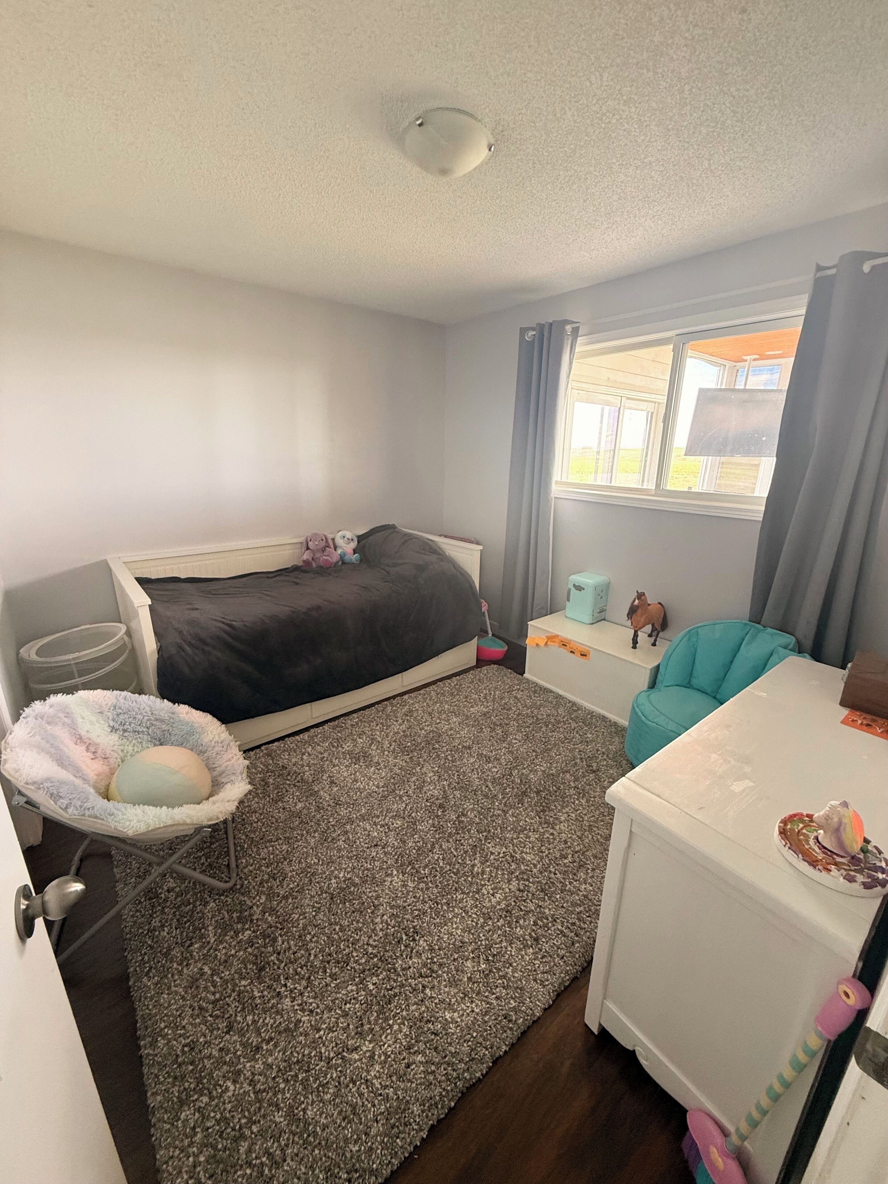 Une chambre à coucher avec un lit, un petit bureau, une chaise, une fenêtre avec des rideaux et un luminaire au plafond.