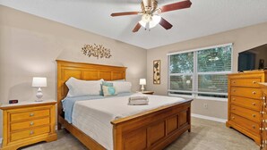 Une chambre à coucher avec un lit en bois, des tables de chevet, une commode, un ventilateur de plafond et une fenêtre avec des stores.