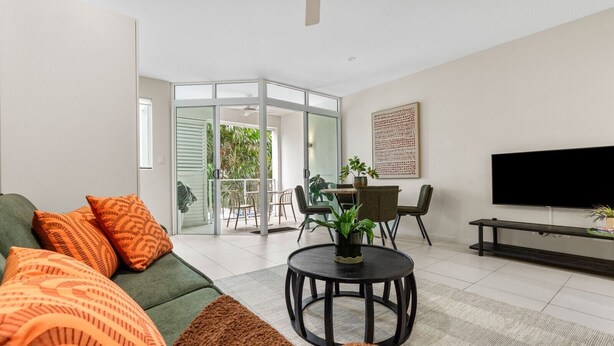 A modern living room with a green sofa, orange pillows, a round coffee table, a TV, and a balcony with outdoor furniture. - Modern Cairns Apartment with Shared Pool, Parking and Close to Esplanade (Cairns City)