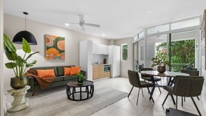 A modern living room with a sofa, dining table, and chairs, a kitchen area, and large windows with a view of greenery. - Modern Cairns Apartment with Shared Pool, Parking and Close to Esplanade (Cairns City)