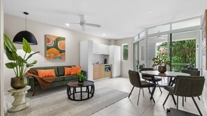 Ein modernes Wohnzimmer mit einer Couch, einem Esstisch und Stühlen, einer Küchenzeile und großen Fenstern mit Blick ins Grüne.