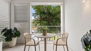 A balcony with a round table and two chairs, a potted plant, and a view of palm trees and parked cars. - Modern Cairns Apartment with Shared Pool, Parking and Close to Esplanade (Cairns City)