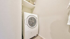 A compact laundry area with a washing machine, a shelf with a basket and a bottle, and a tiled floor. - Grand STAY Hakata Nishinakasu (Fukuoka)