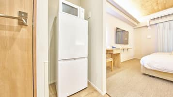 A hotel room with a wooden door, a white refrigerator, a wooden desk, a mirror, a bed, and a textured carpet.