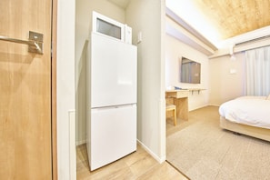 Una habitación de hotel con puerta de madera, refrigerador blanco, escritorio de madera, espejo, cama y alfombra texturizada.