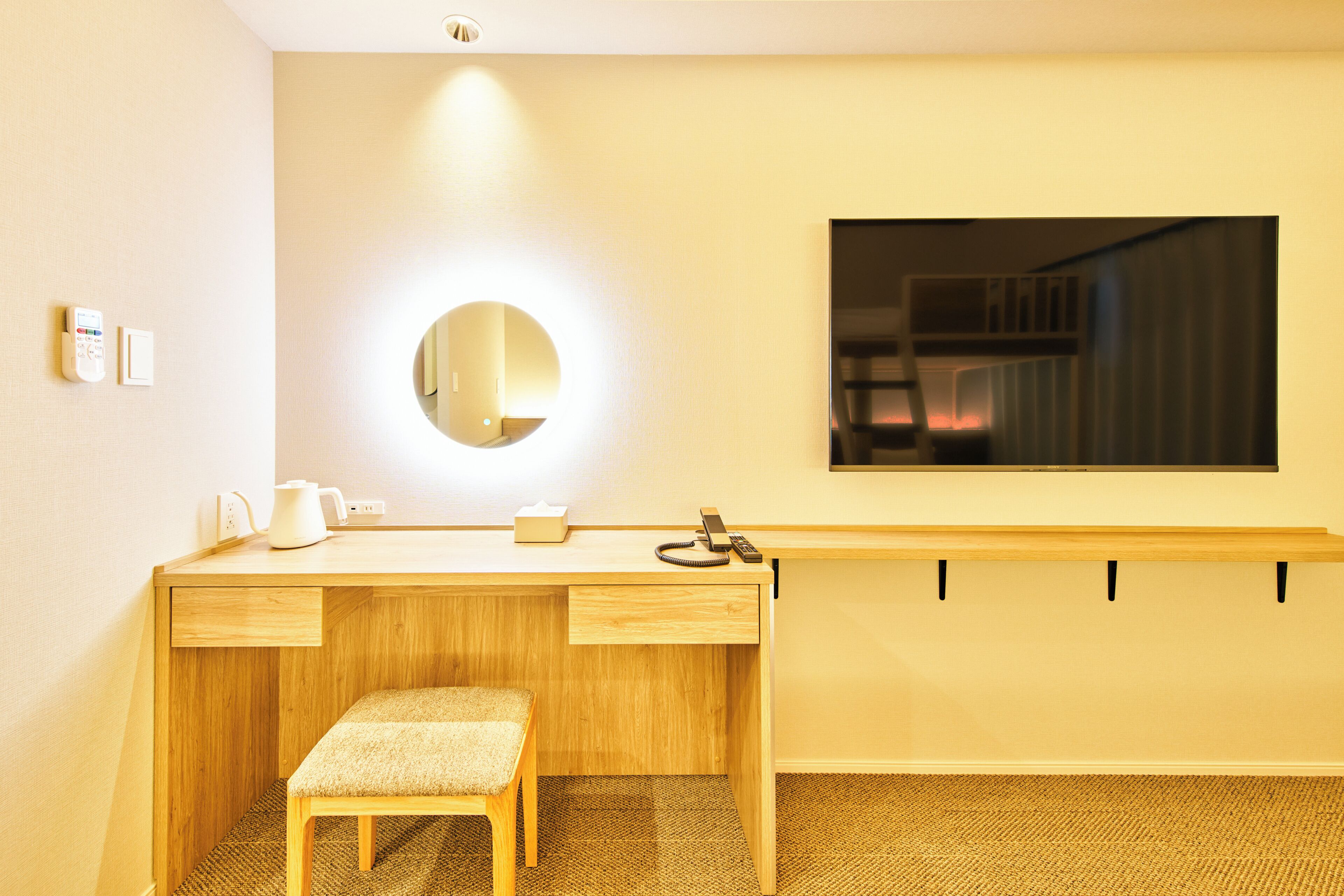 A hotel room with a wooden desk, a small stool, a round mirror, a bedside table, and a wall-mounted TV.