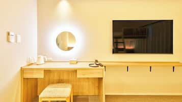 Een hotelkamer met een houten bureau, een klein krukje, een ronde spiegel, een nachtkastje en een aan de muur bevestigde televisie.