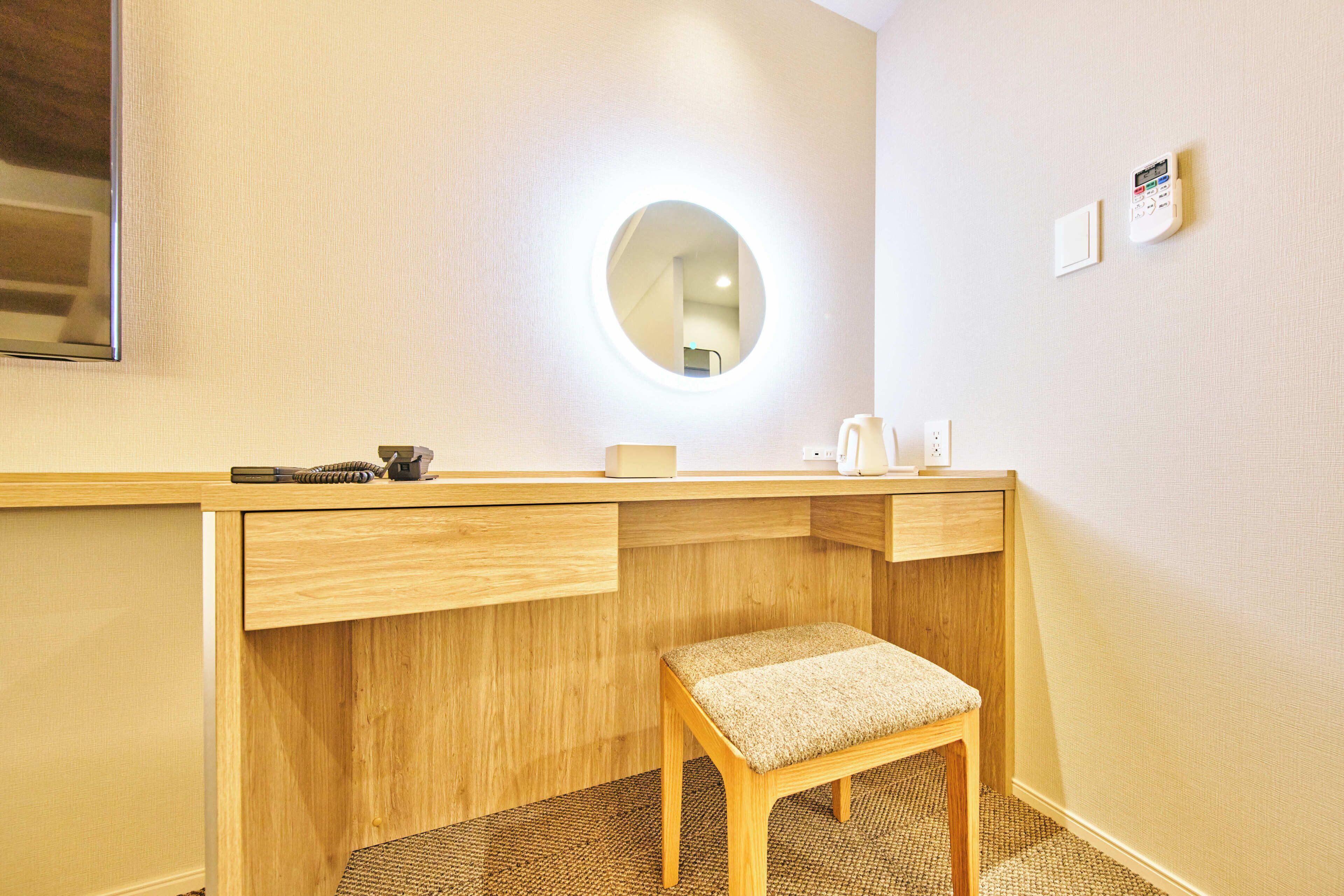 A hotel room with a wooden desk, a round mirror, a small stool, and a wall-mounted thermostat.