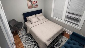 Quarto com cama, uma cadeira, janela com persianas, luminária no teto e um quadro na parede.