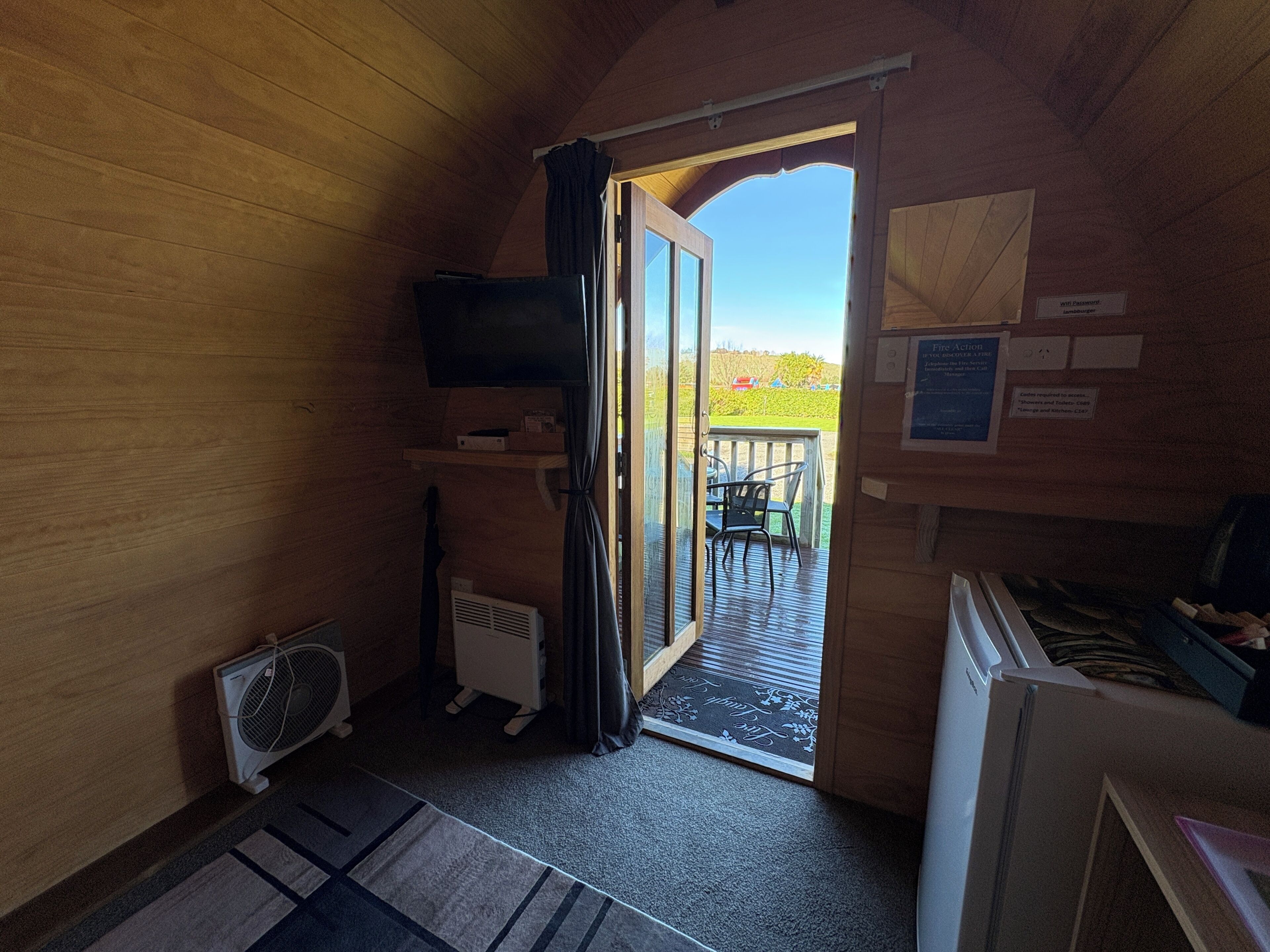A room with a wooden interior, a television mounted on the wall, a small table with chairs, and a door leading to an outdoor area with a view of the sky and greenery.