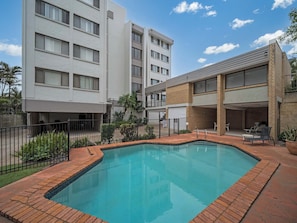 A swimming pool with a brick border, surrounded by a fence and a building with multiple windows and balconies.