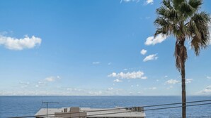 A coastal view with a palm tree, a building with a flat roof, and the ocean in the background. - Entire Apartment: Direct Ocean Views, Pool (Kings Beach)