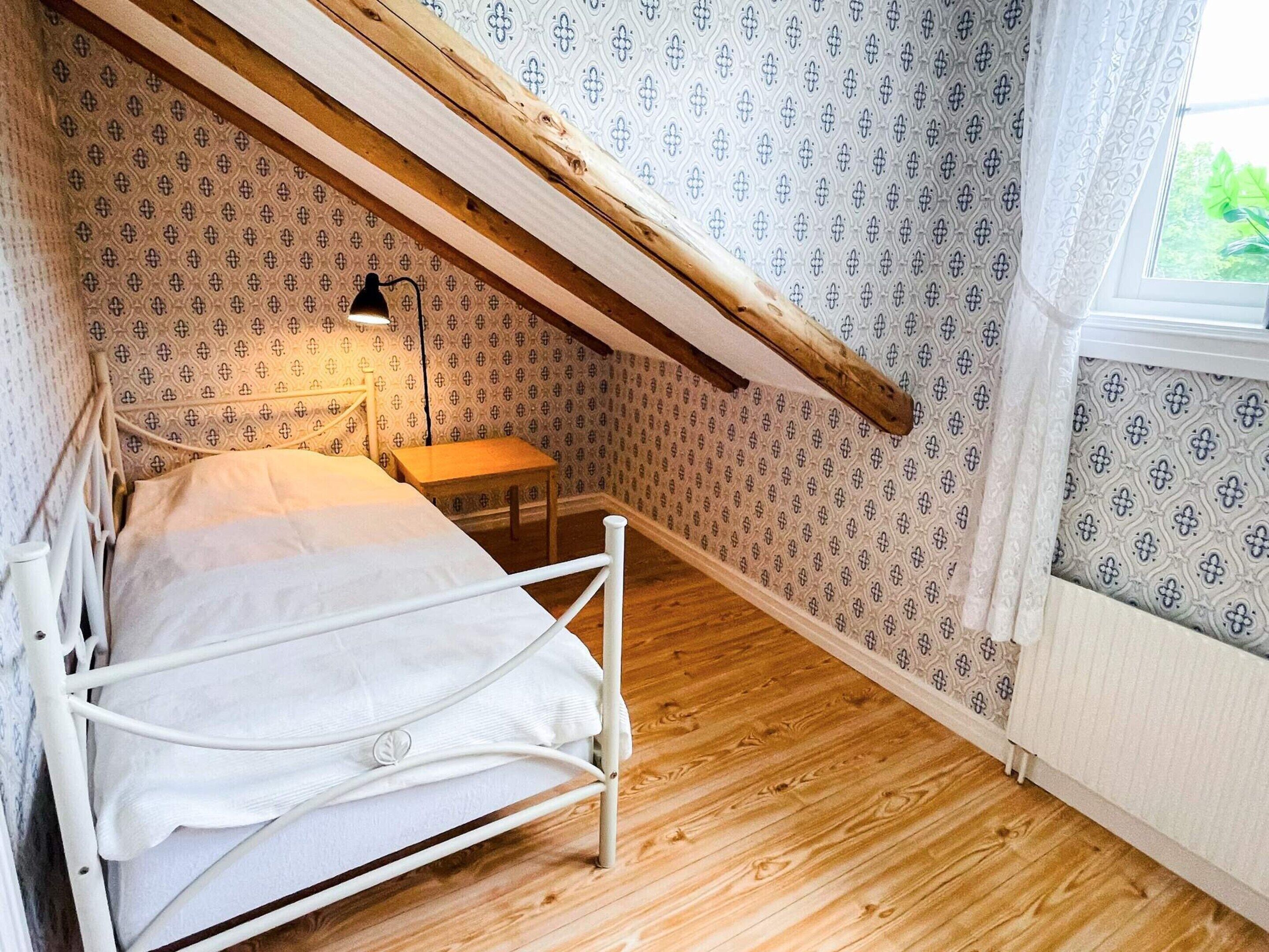 Et soveværelse med skråvæg, seng, natbord, lampe og tapet med mønster.