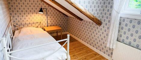 Et soveværelse med skråvæg, seng, natbord, lampe og tapet med mønster.
