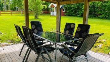 A covered outdoor seating area with a glass table and chairs, surrounded by a wooden deck and a grassy lawn.