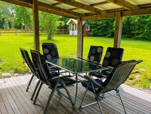 A covered outdoor seating area with a glass table and chairs, surrounded by a wooden deck and a grassy lawn. - 9 Person Holiday Home in Almhult (Almhult)