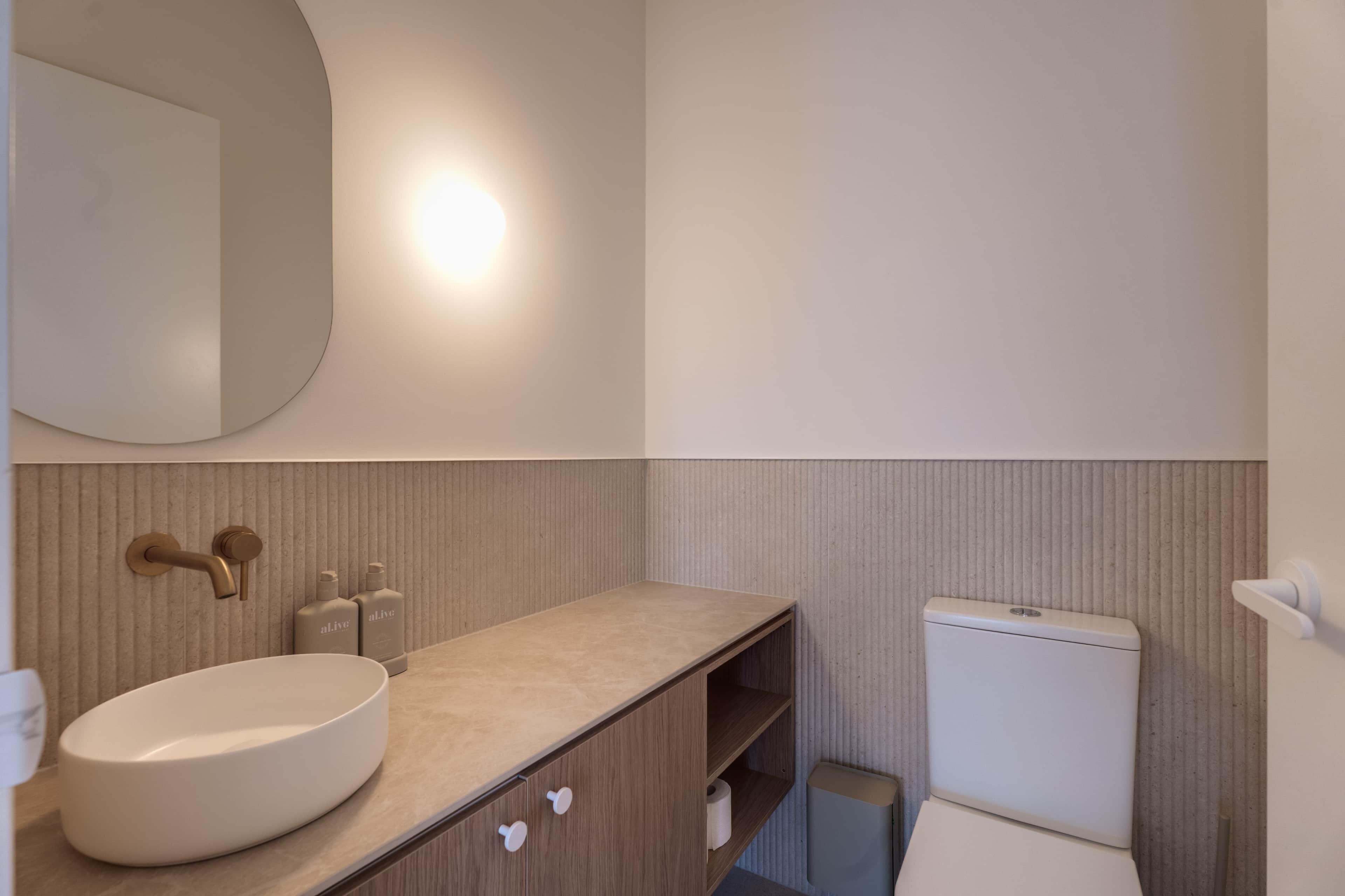 Un baño con un lavamanos redondo, un espejo, un inodoro y un estante con artículos de aseo.