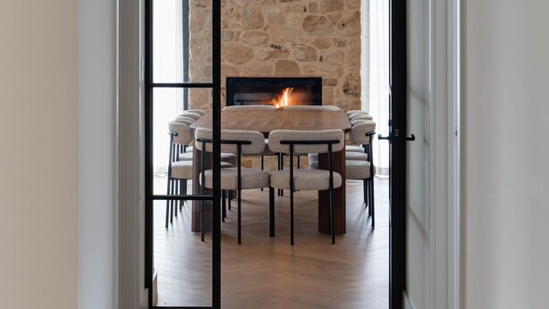 Un couloir mène à une salle à manger agrémentée d’un mur en pierre, d’une cheminée et d’une table en bois avec des chaises.