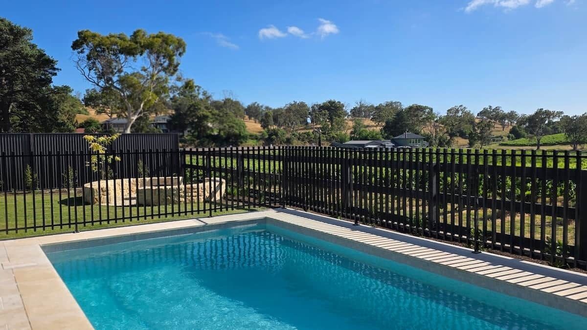 A rectangular swimming pool with a black fence surrounding it, set against a backdrop of trees and hills.