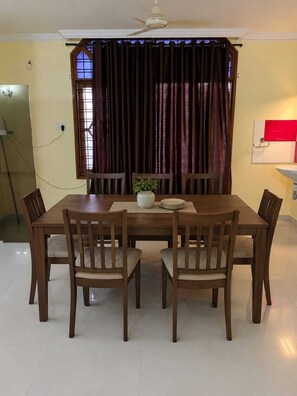 A dining area with a wooden table and chairs, a ceiling fan, a window with curtains, and a wall-mounted light fixture. - Home roots service apartment provide comfort zone long staying client (Hyderabad)