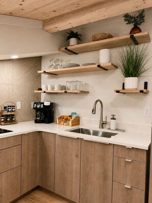 A modern kitchen with wooden cabinets, a stainless steel sink, and multiple shelves with kitchenware and plants.