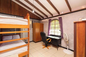 Ein Zimmer mit einem Etagenbett, einem Schreibtisch mit Stuhl, einem Standventilator und einem Fenster mit Vorhängen.