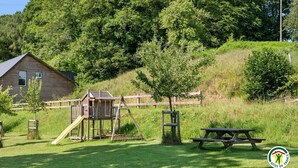 Eine Holzhütte, ein Spielplatz und ein Picknicktisch auf einer grünen Wiese mit Bäumen.