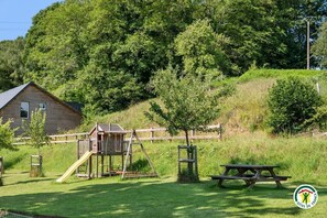 A wooden cabin, a playground, and a picnic table in a grassy area with trees.