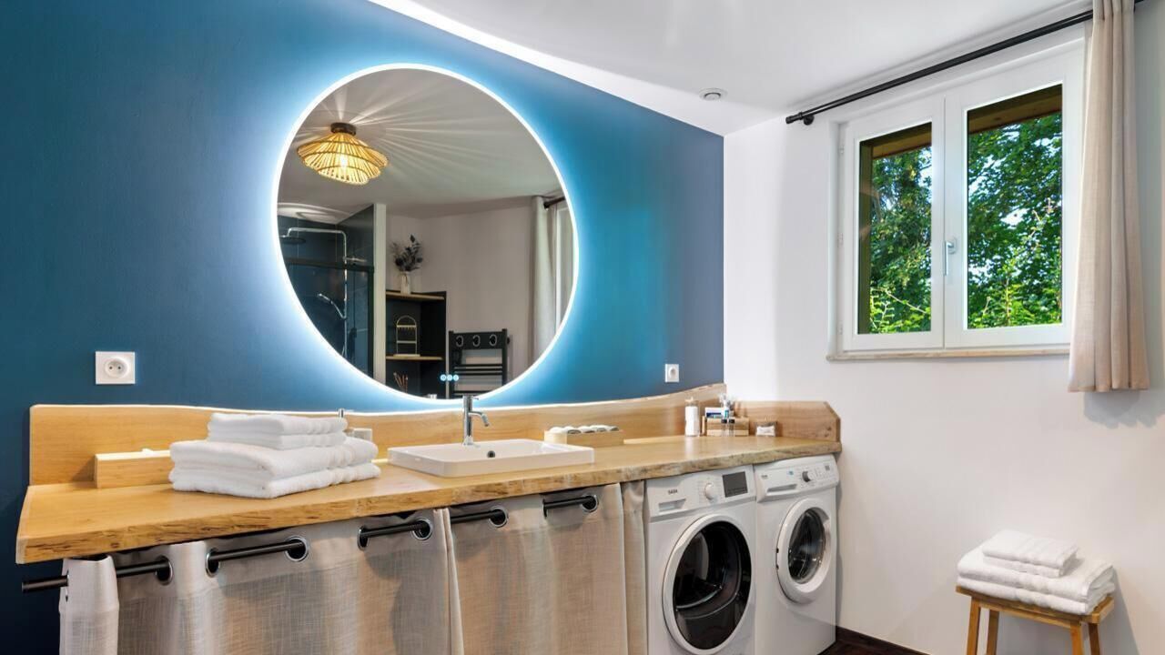Un baño moderno con un espejo grande, un lavamanos, una lavadora y una ventana con vista a árboles.