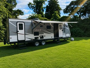 Un camping-car Coleman garé sur de l’herbe, avec une porte latérale, une petite fenêtre et une échelle.