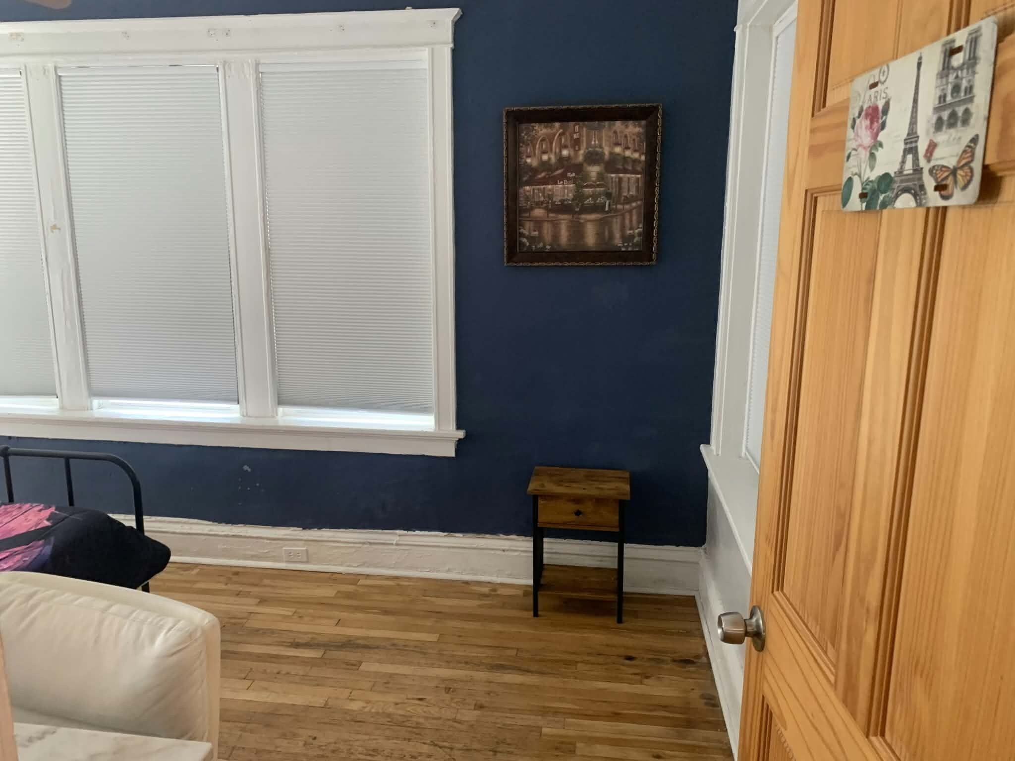 A room with a large window, a small wooden table, a bed with a pillow, and a door.
