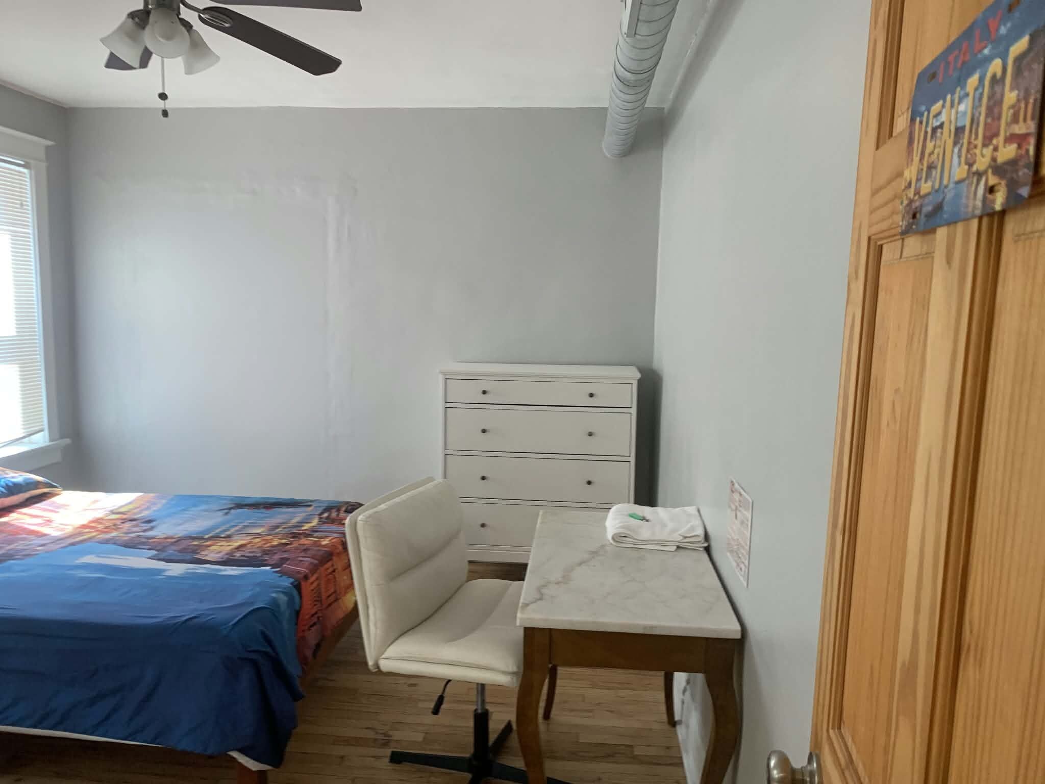 A bedroom with a bed, a desk, a chair, a dresser, and a ceiling fan.