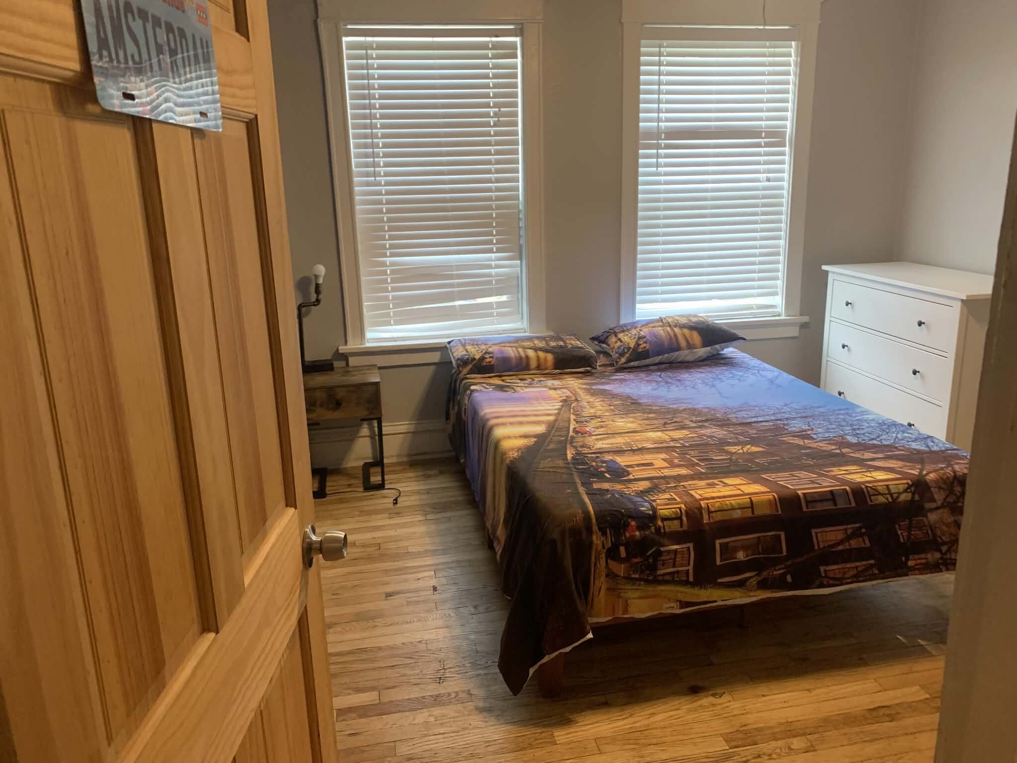 A bedroom with a bed, two windows with blinds, a dresser, and a door.