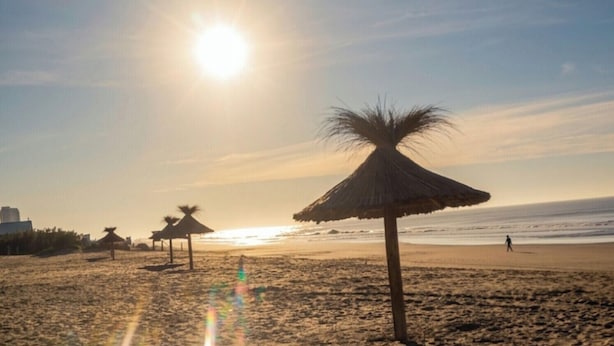 A beach with a sunlit sky, a thatched umbrella, and a person walking on the sand.