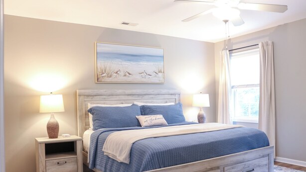 A bedroom with a bed, two bedside tables, a ceiling fan, a window with curtains, and a painting on the wall. - Family Beach Retreat,  Arcade, Fire Pit, Dog Friendly (Surf City)