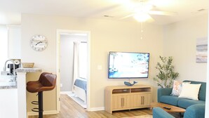 A modern living room with a TV, a wooden cabinet, a blue sofa, a wooden floor, and a ceiling fan. - Family Beach Retreat,  Arcade, Fire Pit, Dog Friendly (Surf City)