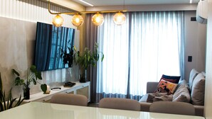 A modern living room with a dining area, a flat-screen TV, and a large window with curtains. - Couple's Apartment with Sea View in Ponta Negra - EFM404 (Natal)
