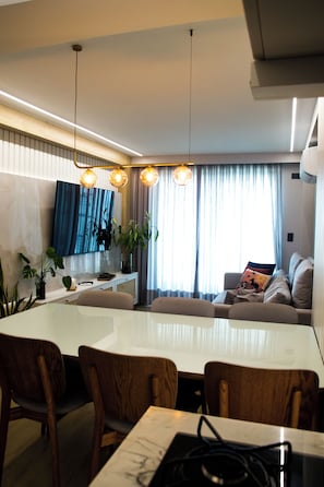 A modern living room with a dining area, a flat-screen TV, and a large window with curtains. - Couple's Apartment with Sea View in Ponta Negra - EFM404 (Natal)