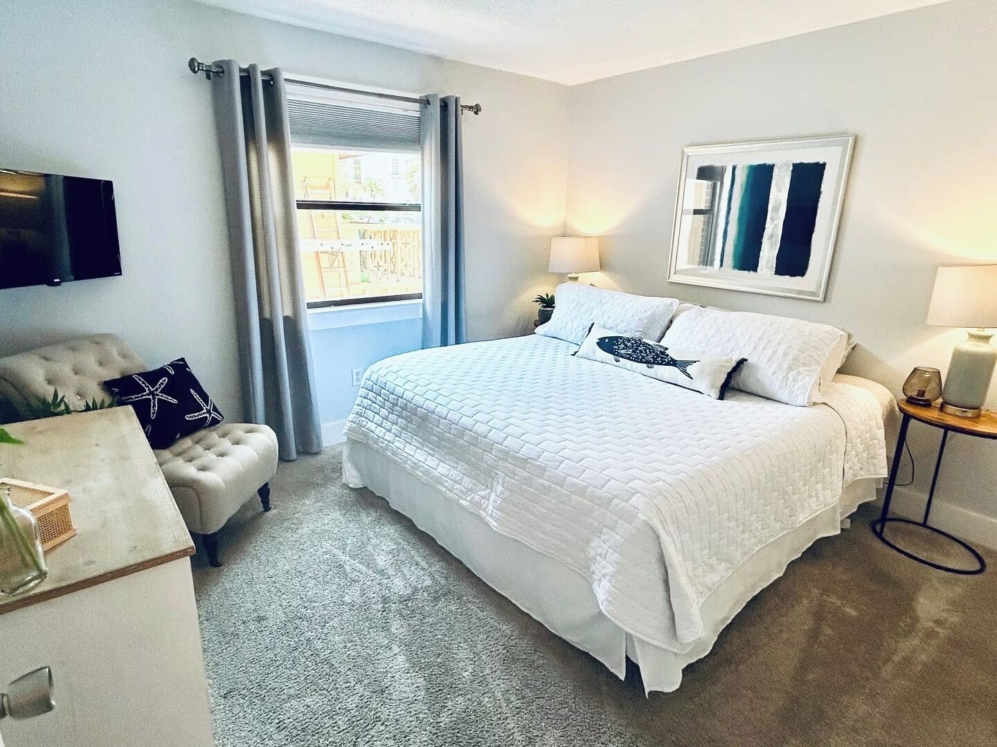 Une chambre à coucher comprenant un lit, un téléviseur, une fenêtre avec des rideaux, une table de chevet et une œuvre d’art accrochée au mur.