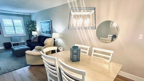 A dining area with a wooden table and white chairs, a hanging light fixture, and a round mirror on the wall. - Venus 205 | Beach Service Included | Beach Getaway (Fort Walton Beach)