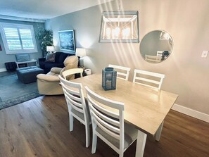 A dining area with a wooden table and white chairs, a hanging light fixture, and a round mirror on the wall. - Venus 205 | Beach Service Included | Beach Getaway (Fort Walton Beach)