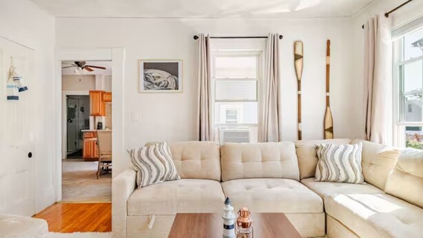 A modern living room with a beige sectional sofa, a wooden coffee table, and a view of the city through large windows. - The Thomas Honeycomb Inn w/ Tiki Bar and Roof Deck (Salem)