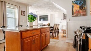 A kitchen with wooden cabinets, a central island, a dining area with chairs, and a painting on the wall. - The Thomas Honeycomb Inn w/ Tiki Bar and Roof Deck (Salem)