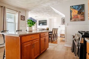 A kitchen with wooden cabinets, a central island, a dining area with chairs, and a painting on the wall. - The Thomas Honeycomb Inn w/ Tiki Bar and Roof Deck (Salem)