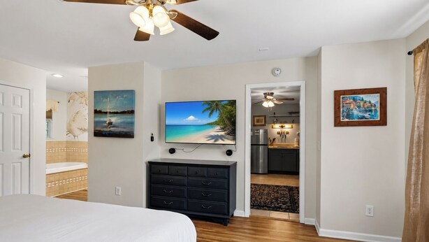 A bedroom with a bed, a TV, a ceiling fan, and a view into the kitchen. - Brick House By the Bay | Bay Walk + Local Life (Shalimar)