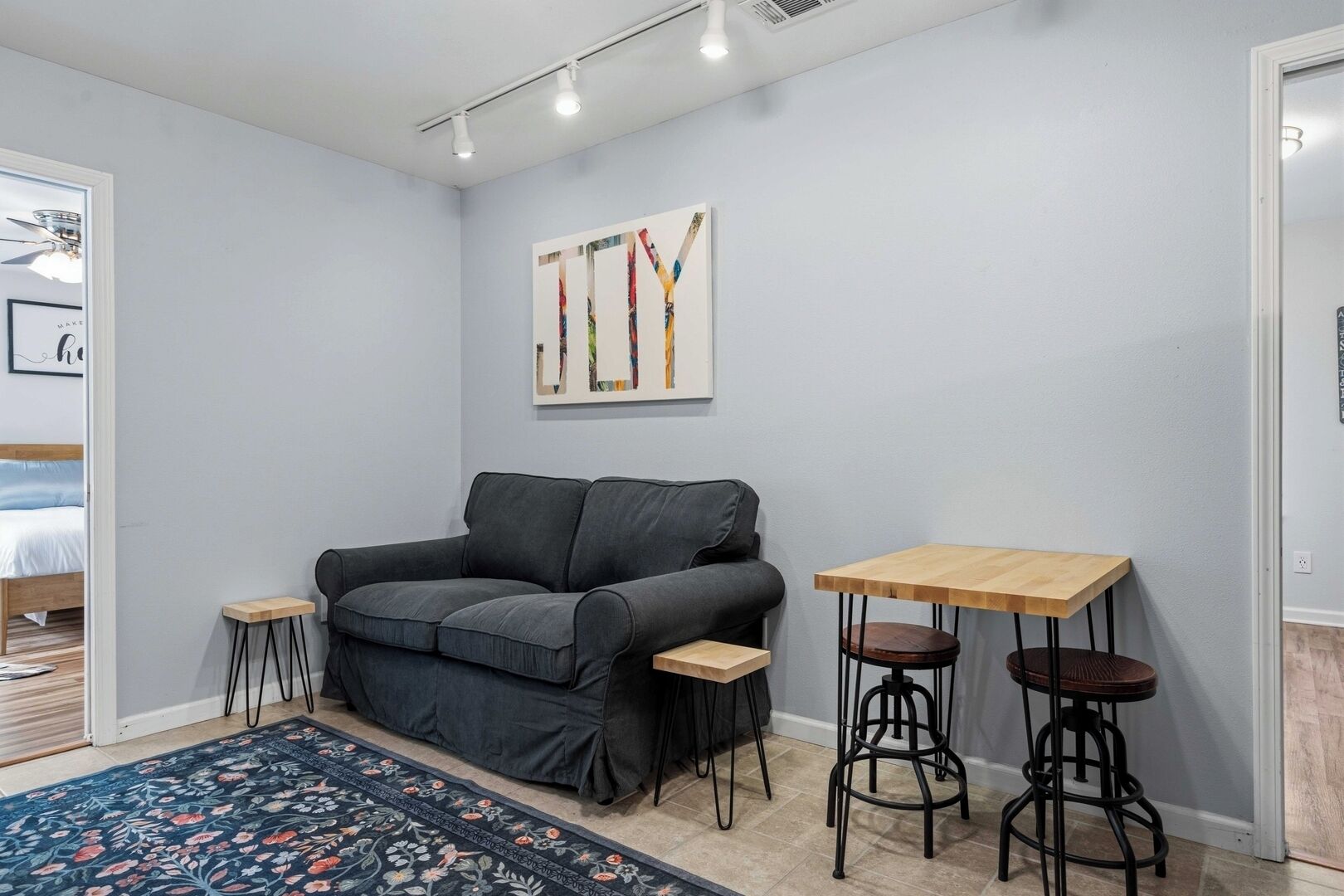 A room with a grey sofa, a small wooden table, two stools, a colorful rug, and a framed abstract painting on the wall.