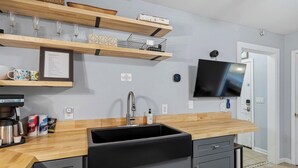 A modern kitchen with a wooden countertop, black sink, and built-in cabinets. There are shelves above the counter holding decorative items and a flat-screen TV mounted on the wall. - Brick House By the Bay | Bay Walk + Local Life (Shalimar)