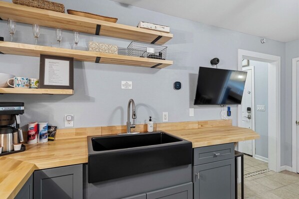 Une cuisine moderne avec un plan de travail en bois, un évier noir et des placards intégrés. Des étagères au-dessus du plan de travail accueillent des objets décoratifs, et un téléviseur à écran plat est fixé au mur.