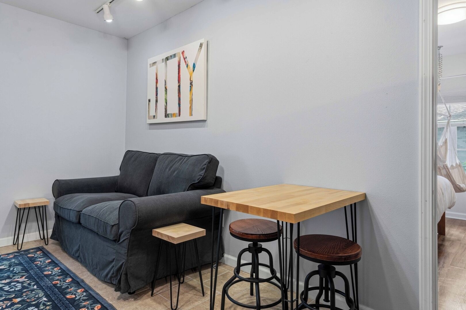 A small living area with a sofa, a wooden table, two stools, a rug, and a wall-mounted artwork.