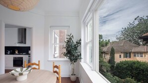 Een kamer met een eettafel, stoelen, een potplant en uitzicht op een huis door een raam.